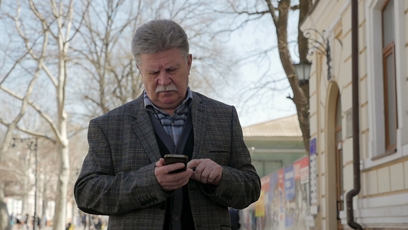 Grey-Headed Man with Mustache Walks and Browses the Net on a Phone in Spring