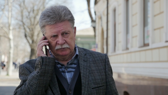 Elderly Man with Mustache Goes and Talks on His Phone in Spring in