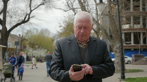 Elderly Man Goes Along a City and Touches His Mobile in Spring in