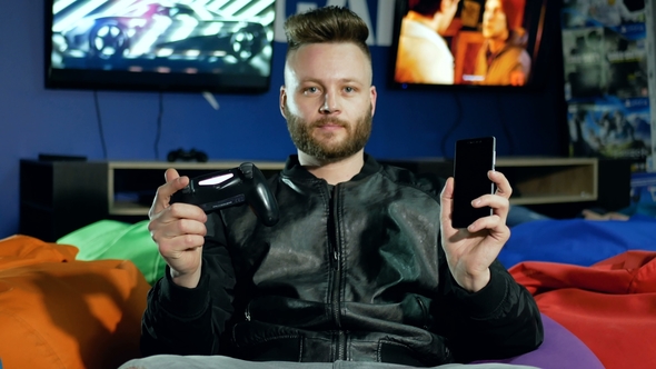 Young Man with a Beard Holds a Smartphone and a Joystick in His Hands