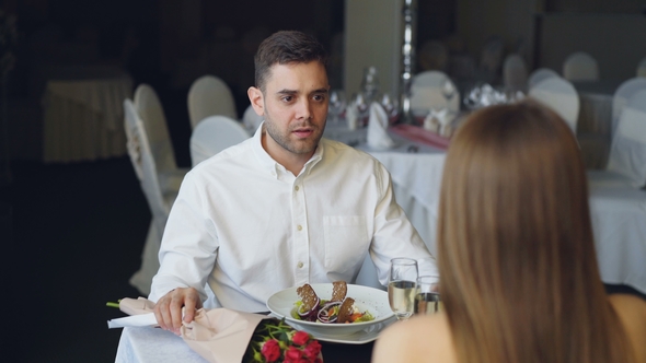 Young Couple Is Quarreling While Dining in Restaurant, Shouting and Gesturing. Bouquet of Roses alt