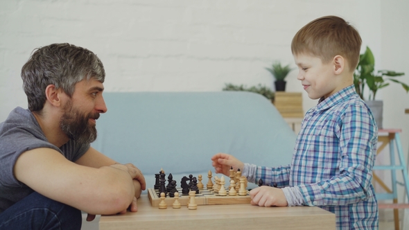 Side View of Father and Son Playing Chess at Home, Talking and Laughing. Chessboard alt