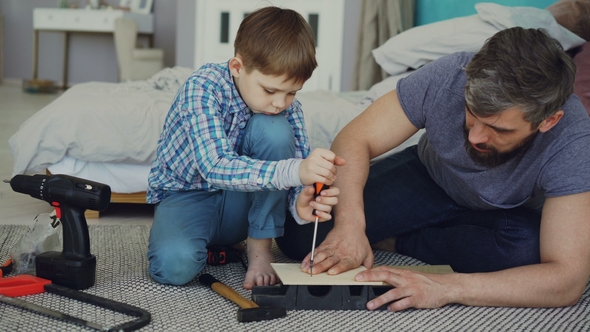 Curious Child Is Driving Screw in Piece of Wood with Screwdriver and His Dad Is Holding Wooden Sheet alt