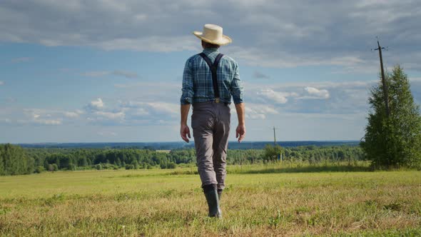 Farmer Wearing Straw Hat Suspenders and Rubber Boots Walking in Mown Field alt