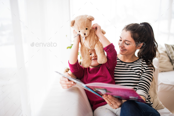 Young mother and a small girl with teddy bear at home, reading a book. Stock Photo by halfpoint