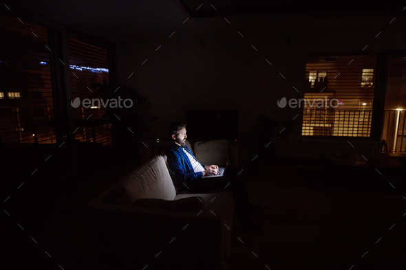 A man working on a laptop at home at night. Stock Photo by halfpoint