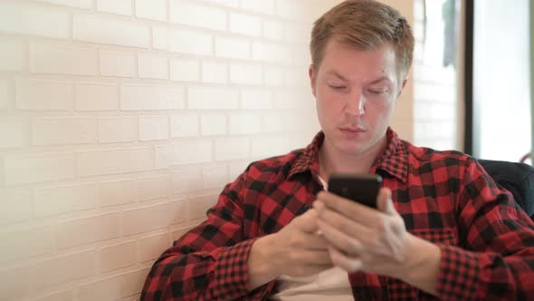 Young Thoughtful Hipster Man Smiling While Using Phone At The Coffee Shop alt