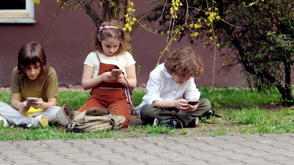 Schoolchildren Sit Down on the Grass After School, Get Their Smartphones Out of Their Satchels and alt