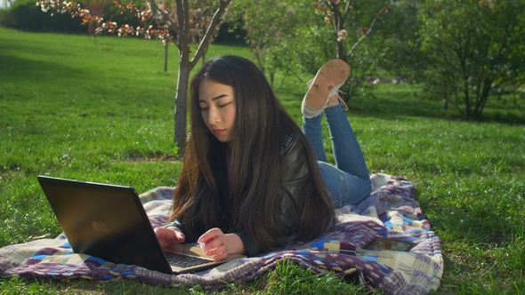 Asian Girl Working with Her Notebook Lying on Lawn alt