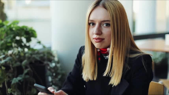 Young Blonde Woman Looks Straight Into the Camera Holding a Smartphone alt