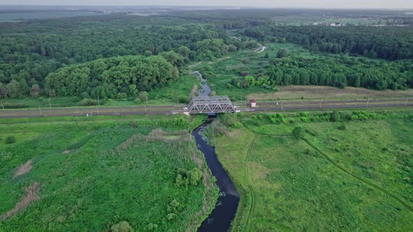 Small River Flows Smoothly Between Green Fields and a Railway Bridge alt