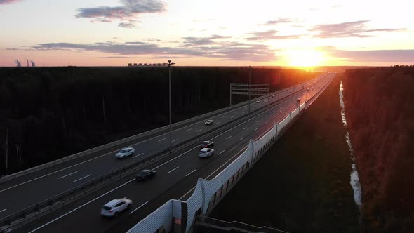 Wide Road Among Forests and Driving Cars at Orange Sunset alt