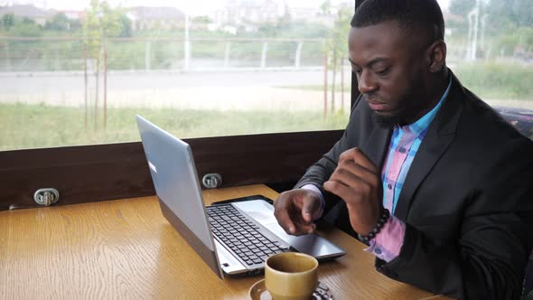 Afro American Businessman is Typing a Message on Laptop and Drinks Cup of Coffee alt