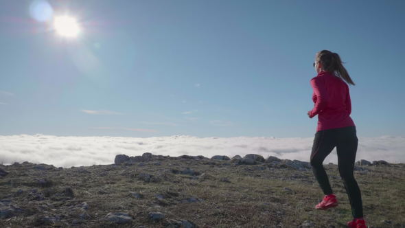 Young Caucasian Woman in Sunglasses Is Jogging in Mountains Above Clouds at Sunny Day. Sky Running alt