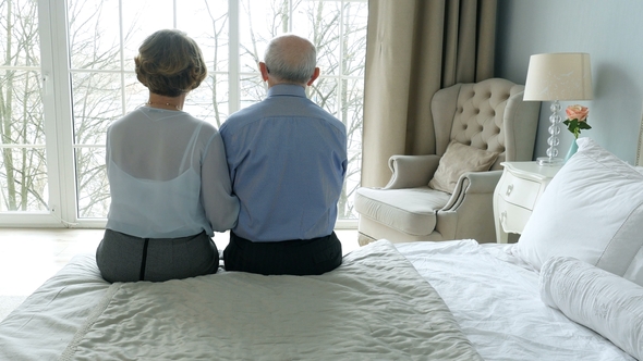 Rear View Lovely Elderly Couple Sitting In Bedroom, Embracing and Looking Out The Window