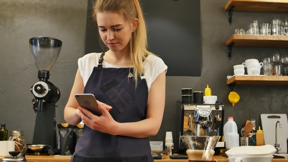 Delighted Female Barista Using Cell Phone at Work Place alt
