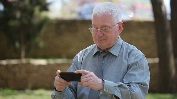 Cheery Old Man Sits and Looks at His Smartphonescreen in a Street on a Sunny Day