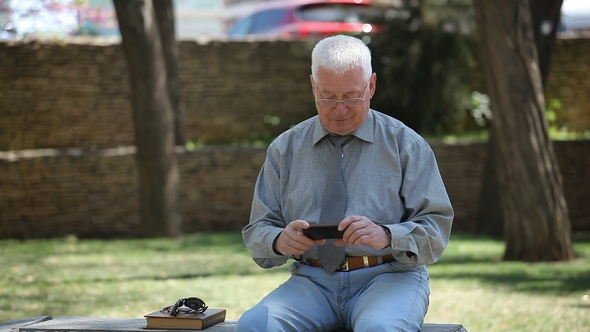 Grey-haired Man Sits and Looks at His Mobile Screen in a Street on a Sunny Day