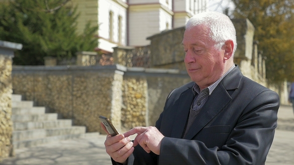 Senior Man Touches the Screen His Phone in a Street in Spring
