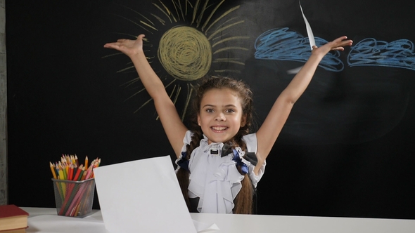 Attractive Girl Sits at a Desk with a Blackboard Behind Her. Happy Schoolgirl Throws alt