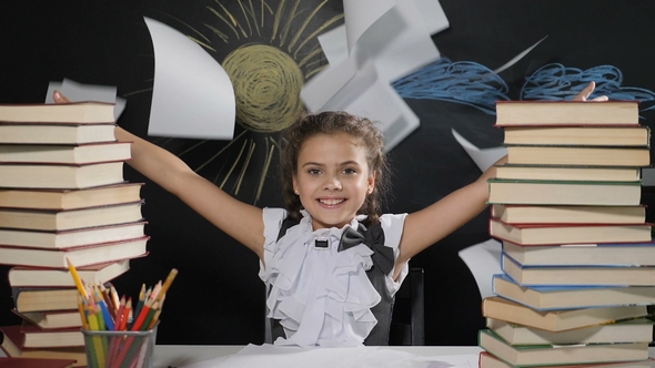 Attractive Girl Sits at a Desk with a Blackboard Behind Her. Happy Schoolgirl Throws alt