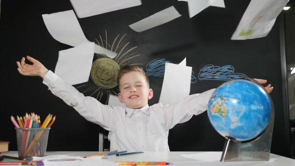 Cute Boy Sits at a Desk with a Blackboard Behind Her. Happy Schoolboy Throws Paper alt
