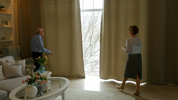 Elderly Couple Opened The Curtains In TThe Living Room, Embracing And Looks Out The Window alt