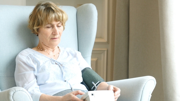 Elderly Woman Sitting in Living Room Near the Window and Measuring Blood Pressure