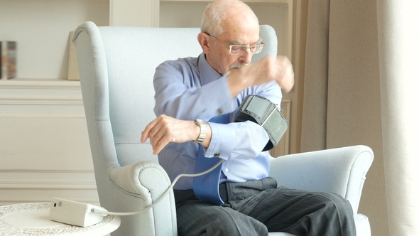 Elderly Man Sitting in Living Room Near the Window and Measuring Blood Pressure