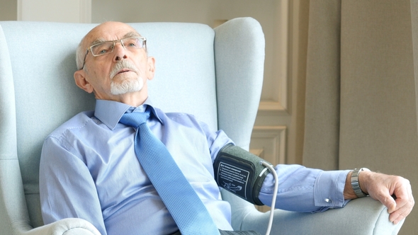 Senior Man Sitting in Living Room Near the Window and Measuring Blood Pressure