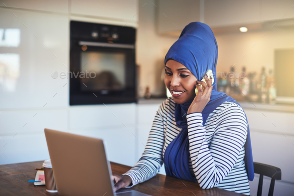 Smiling Arabic entrepreneur talking on a phone in her kitchen - Stock Photo - Images