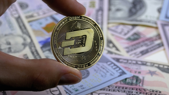 Hand in Fingers Holds a Gold Coin Dash on a Background with Bills of Dollars alt