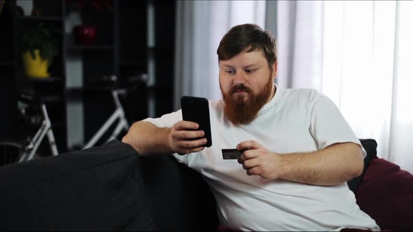 Serious Fat Man Types a Number of His Credit Card in the Smartphone Sitting on the Couch alt