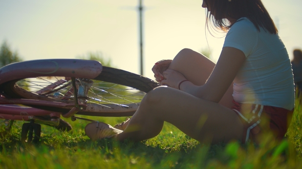 Girl Sitting on the Grass and Scrolls the Wheel of a Bicycle, in the Background Riding Another Girl alt