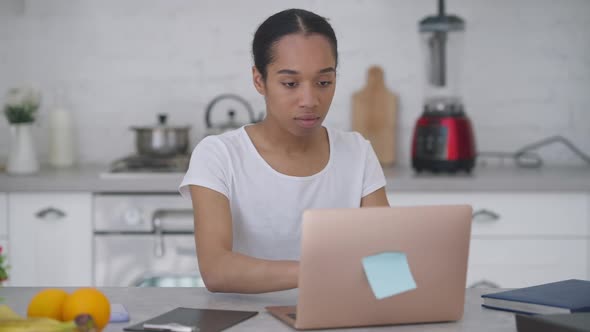 Young Woman Closing Laptop Taking Smartphone and Walking Away From Kitchen Table alt