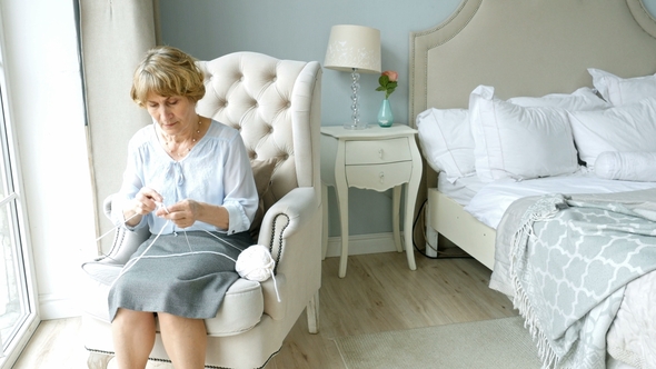 Elderly Woman Knits Sitting at the Window in Bed Room