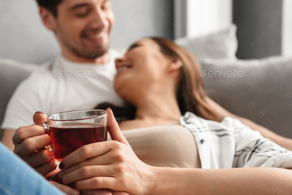 Blurry photo of cheerful couple lying on sofa in flat, while wom