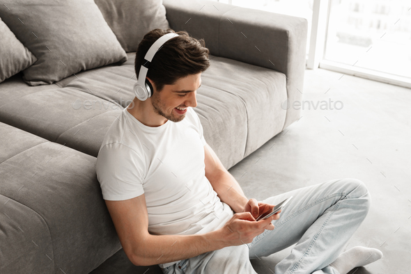 Modern man 30s in white t-shirt sitting on floor indoor, while u Stock Photo by vadymvdrobot