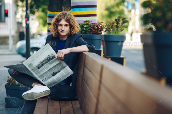 Red haired hipster man sitting on bench with newspaper - Stock Photo - Images