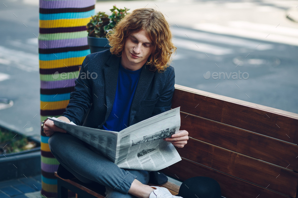 Red haired hipster man sitting on bench reading newspaper - Stock Photo - Images