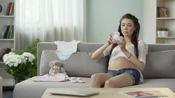 Young Pregnant Woman Taking a Look at Children Clothes on Sofa, Baby Fashion alt
