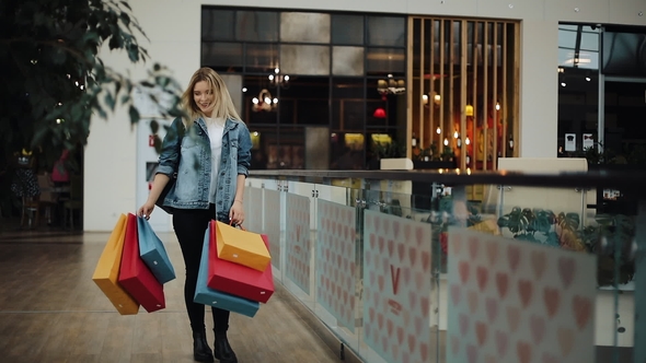Young Blonde Woman in Jeans Jacket Walks Around a Shopping Mall with Colorful Bags alt