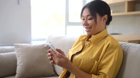 Happy Multiracial Woman Sitting on the Couch and Spending Time Online with Her Smartphone