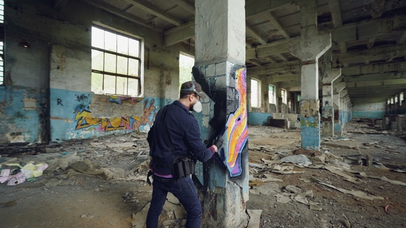 Pan Shot Of Adult Man Graffiti Artist In Protective Face Mask And Gloves Painting On High Pillar By Silverkblack