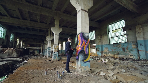 Graffiti Artist in Protective Mask Is Painting on High Column in Abandoned Industrial Building alt