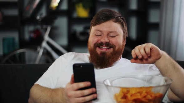 Fat Man Smiles While He Reads Something in His Smartphone and Eats Potato Chips alt