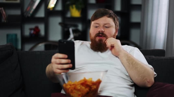 Fat Man Smiles While He Reads Something in His Smartphone and Eats Potato Chips alt