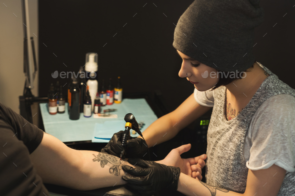 Young woman tattooist doing tattoo on male arm Stock Photo by Prostock-studio