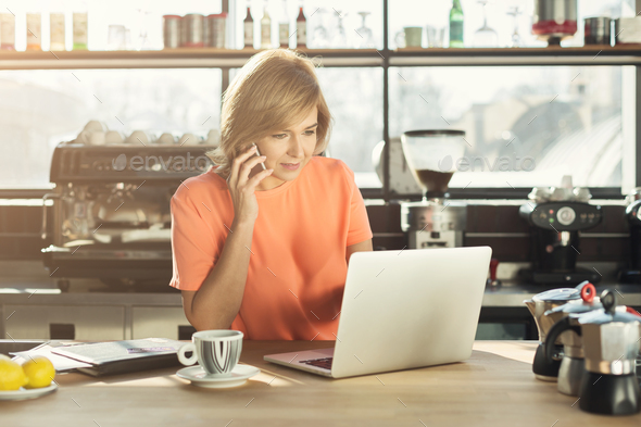 Middle-aged woman barista working on laptop and talking on smartphone - Stock Photo - Images