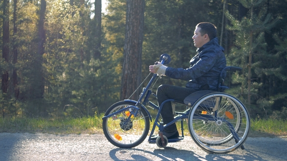 A Handicapped Man Goes on a Wheelchair on a Forest Road, Side View alt